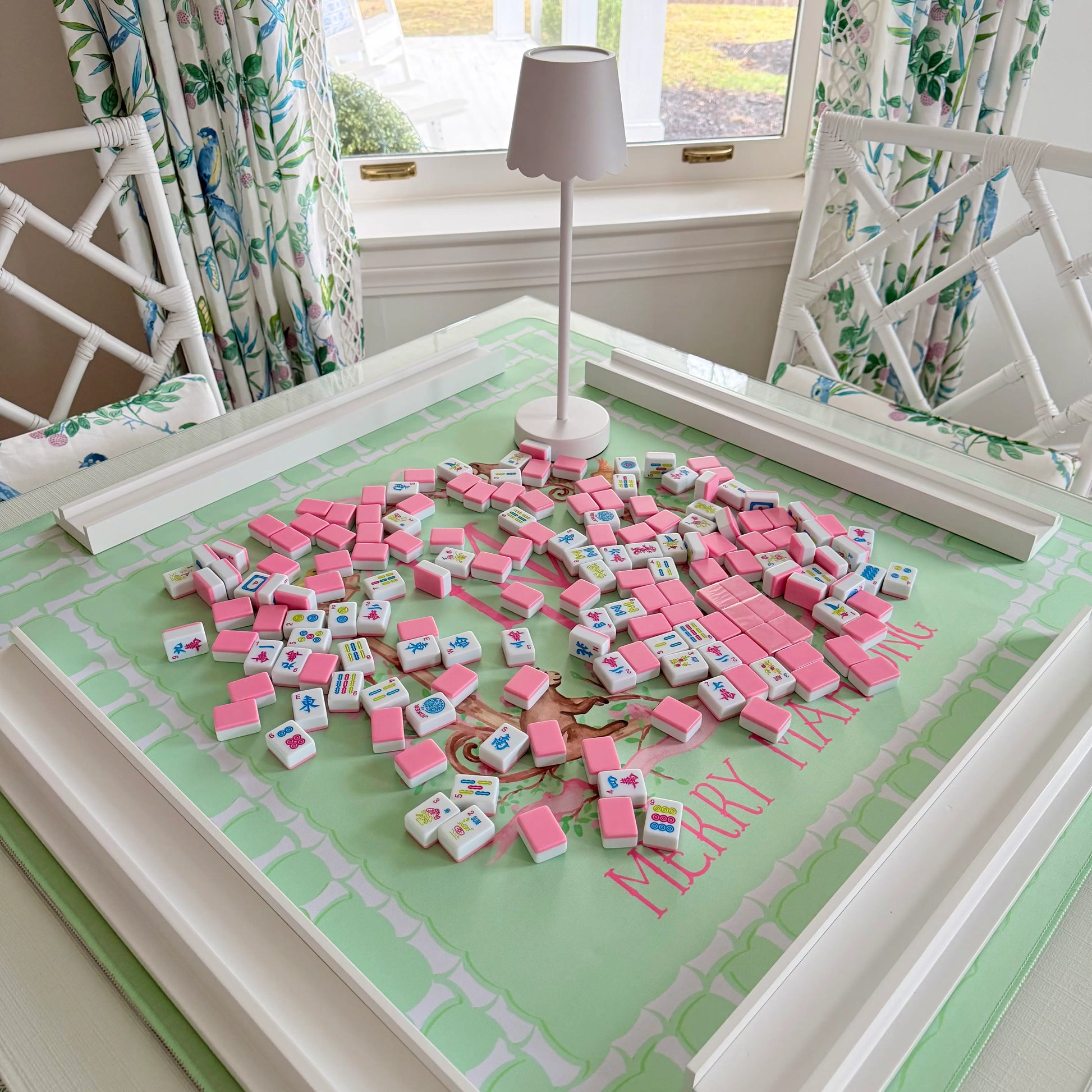Light Green Mahjong Mat with a lattice bamboo border and the TheMerryMahjongCo Logo featuring floral elements alongside monkeys, birds and butterflies. On the mat is the cabana pink tile set, wooden racks and a white LED cordless lamp.