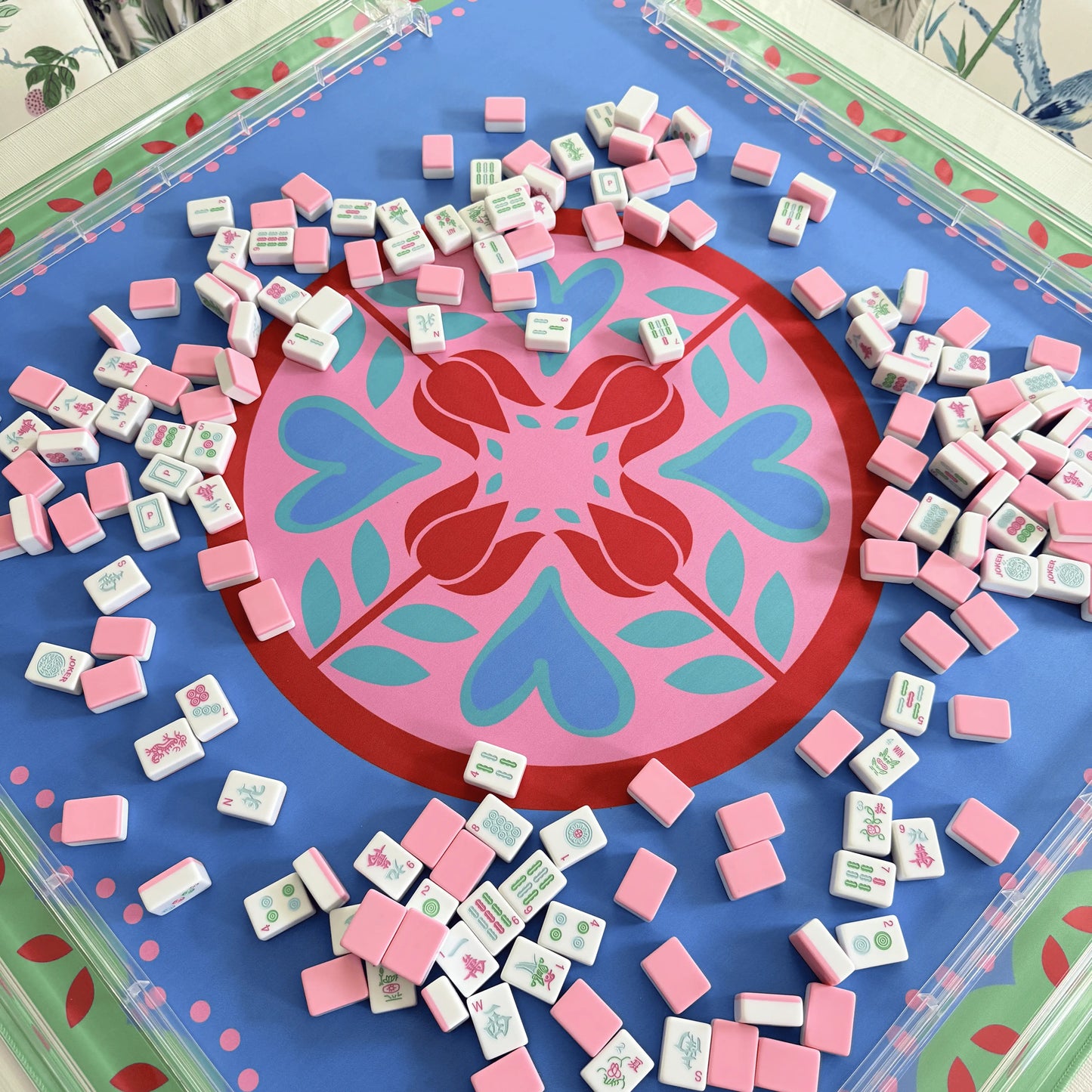 Photograph of cabana pink mahjong tiles spread across a festive Hearts & Flowers mahjong mat featuring pastel azure, green, light magenta, red, and cyan accents, with transparent clear mahjong racks.