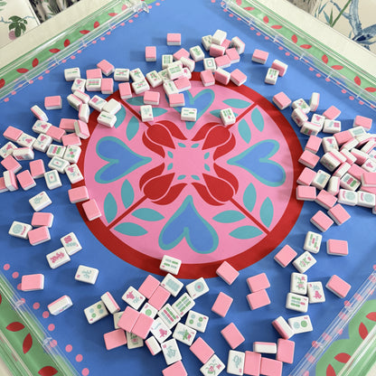 Photograph of cabana pink mahjong tiles spread across a festive Hearts & Flowers mahjong mat featuring pastel azure, green, light magenta, red, and cyan accents, with transparent clear mahjong racks.
