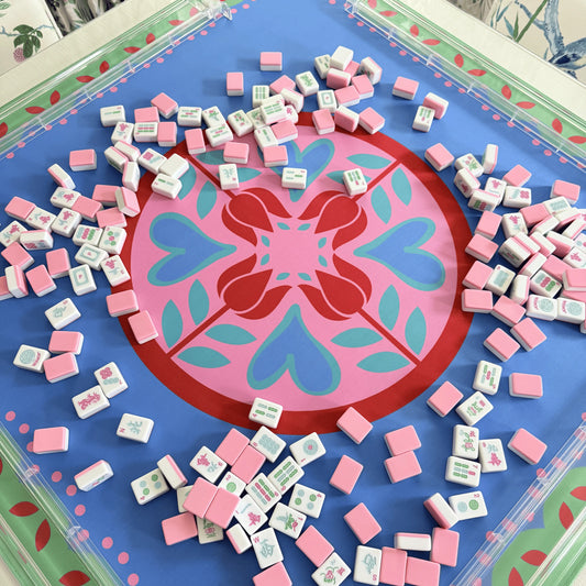 Photograph of cabana pink mahjong tiles spread across a festive Hearts & Flowers mahjong mat featuring pastel azure, green, light magenta, red, and cyan accents, with transparent clear mahjong racks.