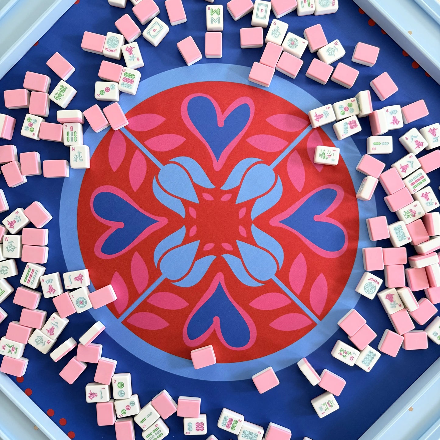 Closeup photograph of cabana pink mahjong tiles arranged around the center of a festive Hearts & Flowers mahjong mat featuring vivid azure and red tones with soft gray green accents, with blue mahjong racks placed along the edges of the photo.