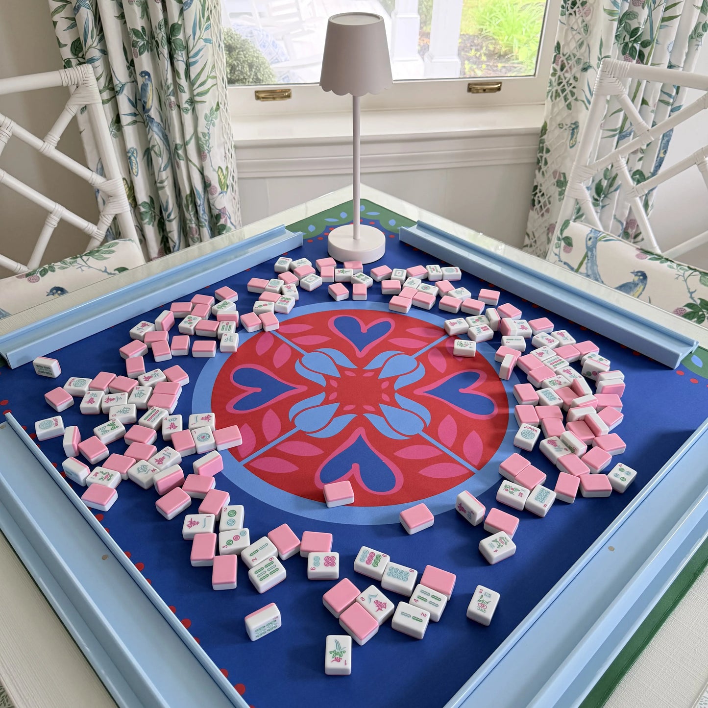 Photograph of cabana pink mahjong tiles spread across a festive Hearts & Flowers mahjong mat featuring vivid azure and red tones with soft gray green accents, with blue mahjong racks along the edges and a cordless white LED lamp placed at the side of the mat.