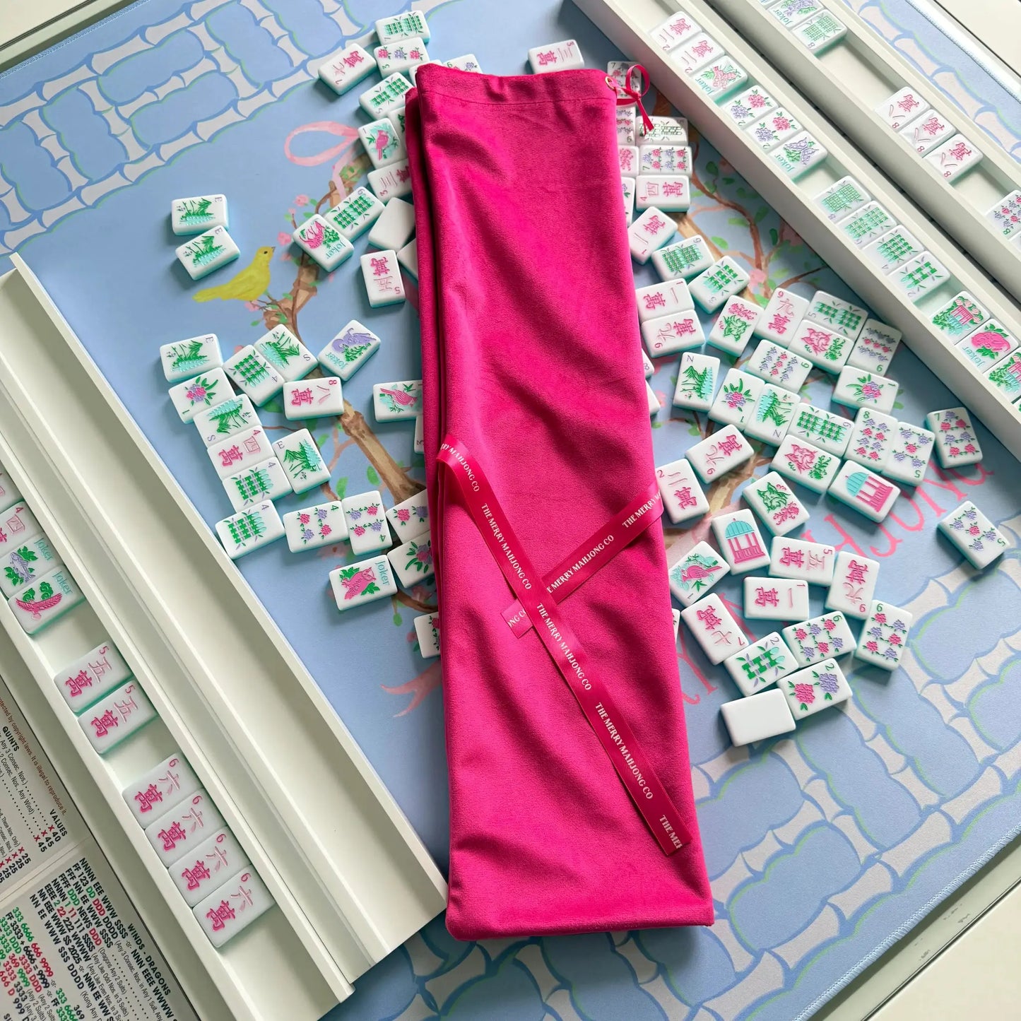 Hot pink velvet mahjong rack bag showcasing its themerrymahjongco ribbons, resting on the blue Bamboo Blossom Mahjong Mat with the Southern Springs Tile set and White Wooden Racks.